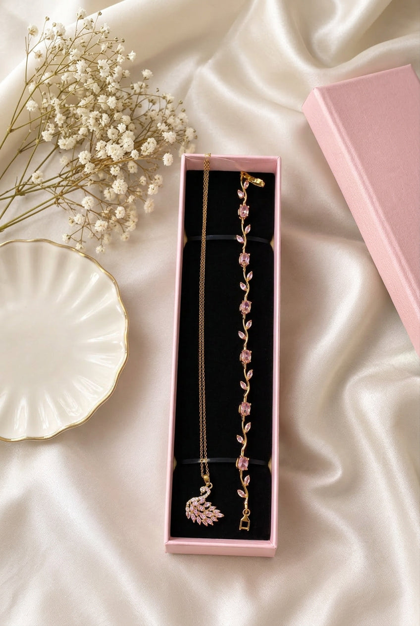 A gold-toned jewelry set with pink gemstone accents, including a long vine bracelet and a matching necklace with a swan pendant, presented in a pink box on a beige satin fabric background.